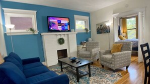 Ein Wohnzimmer mit Kamin, Fernseher, einem blauen Sofa, gemusterten Sesseln und einem Essbereich mit Tisch und Stühlen.