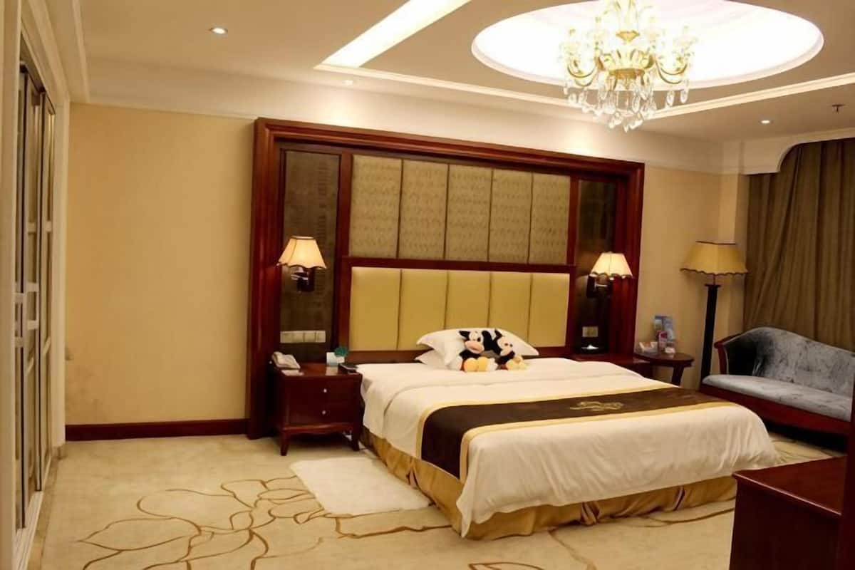 A hotel room with a large bed, bedside lamps, a desk, a chair, and a chandelier.