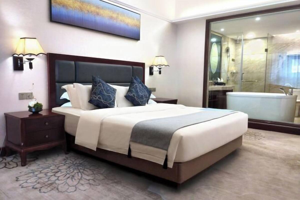 Une chambre d’hôtel avec un grand lit, deux tables de chevet, une commode, une lampe fixée au mur, un tableau et une salle de bain avec une baignoire et une douche.