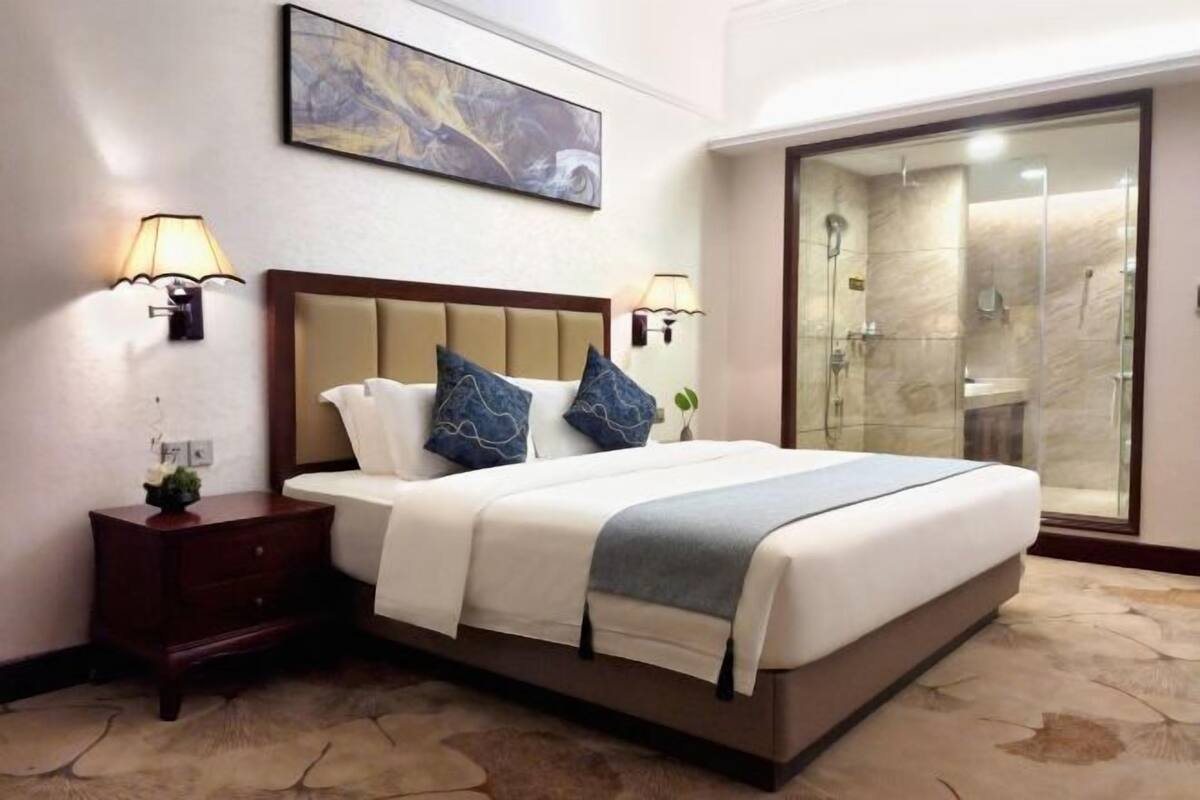 Une chambre d’hôtel avec un grand lit, deux lampes de chevet, une table de nuit, un tableau au mur et une douche en verre.