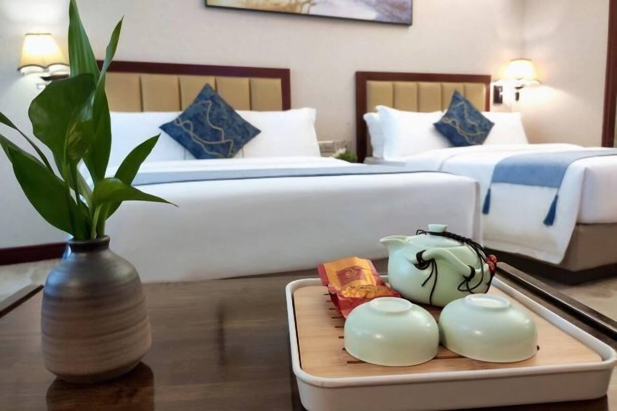 Une chambre d’hôtel avec deux lits, une table de chevet ornée d’un vase et de théières, et une lampe fixée au mur.
