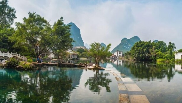 A tranquil water body with a wooden bridge, lush greenery, and distant mountains. - Dong Zheng International Hotel (Hechi)