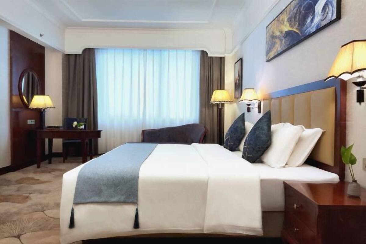 Une chambre d’hôtel avec un lit, des lampes de chevet, un bureau, une chaise, une fenêtre avec des rideaux et un tableau au mur.