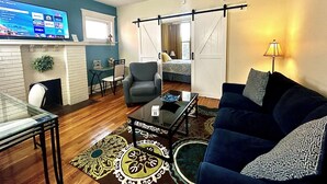 A living room with a sofa, armchair, coffee table, and a TV displaying various channels.