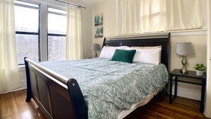 A bedroom with a bed, two bedside tables, a window with curtains, a ceiling fan, and a painting on the wall.