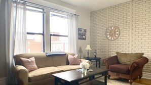 Una sala de estar con un sofá color beige, un sillón marrón, una mesa de centro de madera y un tapete estampado. Hay un reloj en la pared y una ventana con cortinas.
