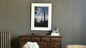 Una habitación con un armario de madera oscura, un jarrón con flores blancas, un cuadro de un puente y un sillón color beige.