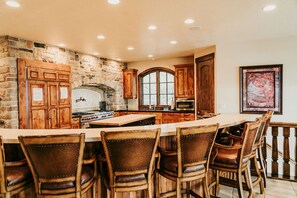 A spacious kitchen with a stone wall, wooden cabinets, a large island, and bar stools. - LAKEFRONT Castle on the Lake | 10BR Sleeps 50 | Private Beach, Hot Tub, Theater & Fire Pit (Fish Haven)