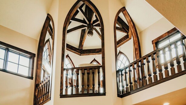 Une chambre spacieuse aménagée dans un grenier, avec un plafond haut, des poutres apparentes et un balcon doté d’une balustrade en fer forgé.