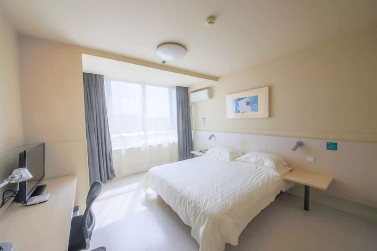 Hotellihuone, jossa on sänky, työpöytä tietokoneella, tuoli, yöpöytä, lamppu ja ikkuna, jossa on verhot.