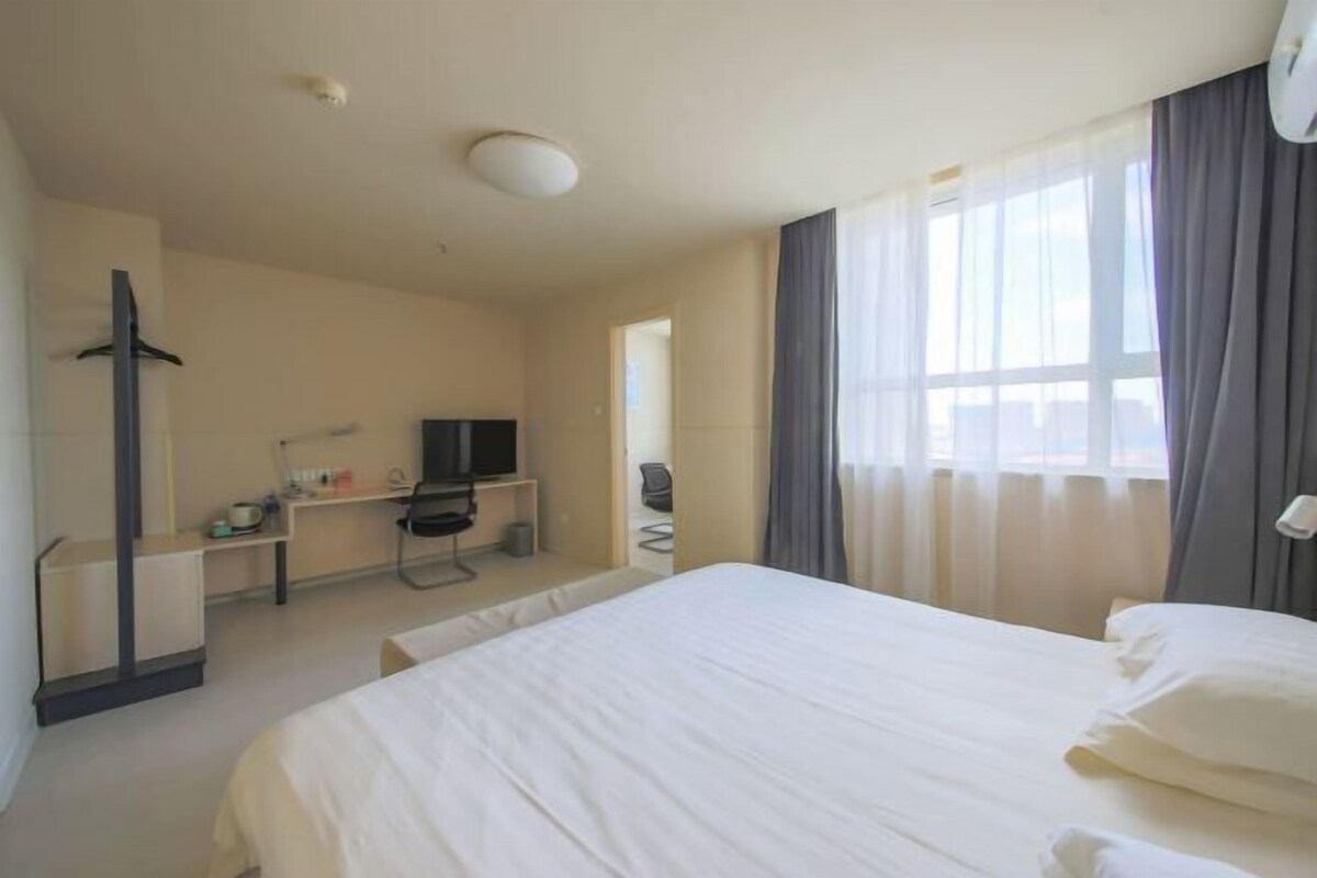Una habitación de hotel con una cama grande, un escritorio con computadora, televisor y ventana con cortinas.