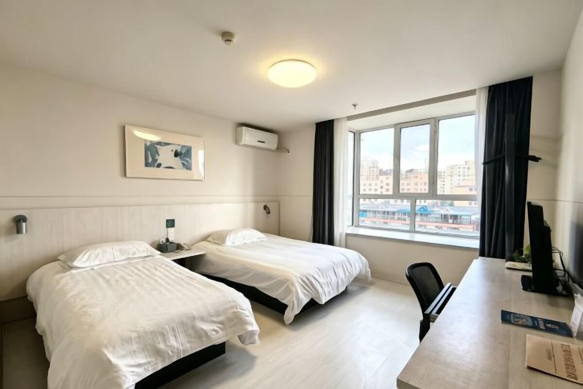 A hotel room with two beds, a desk, a chair, a window with a view of buildings, and a framed picture on the wall.