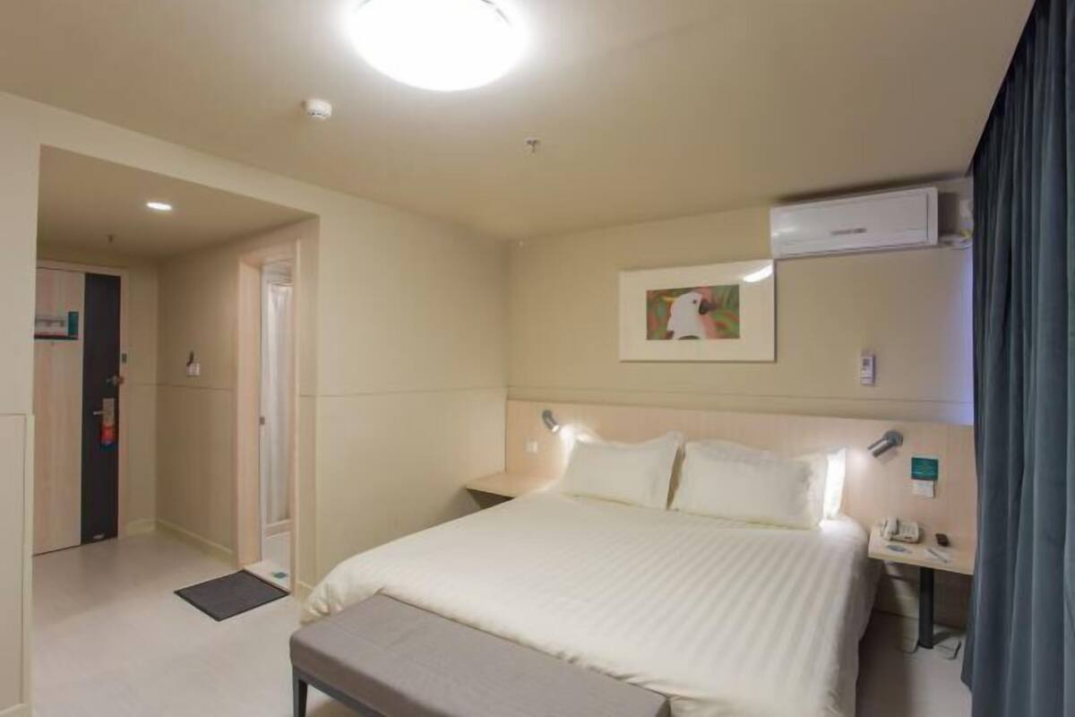 Hotellihuone, jossa on sänky, yöpöytä, lamppu, ilmastointilaite ja taulu seinällä.