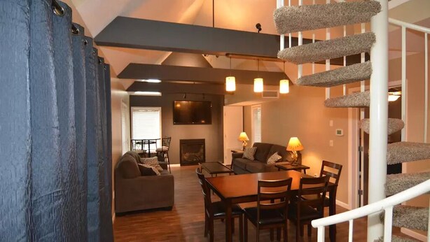 A spacious living room with a wooden dining table, a sofa, and a staircase with a carpeted handrail. - Peek' n Peak, Camelot  Loft Condo,  Clymer, NY, 250 Yards to Chair 8, Sleeps 14 (Clymer)