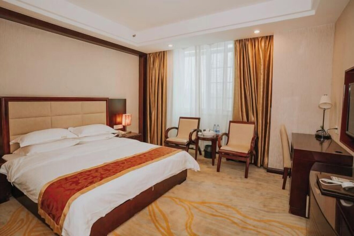 A hotel room with a large bed, a desk, two chairs, a small table, and a window with curtains.