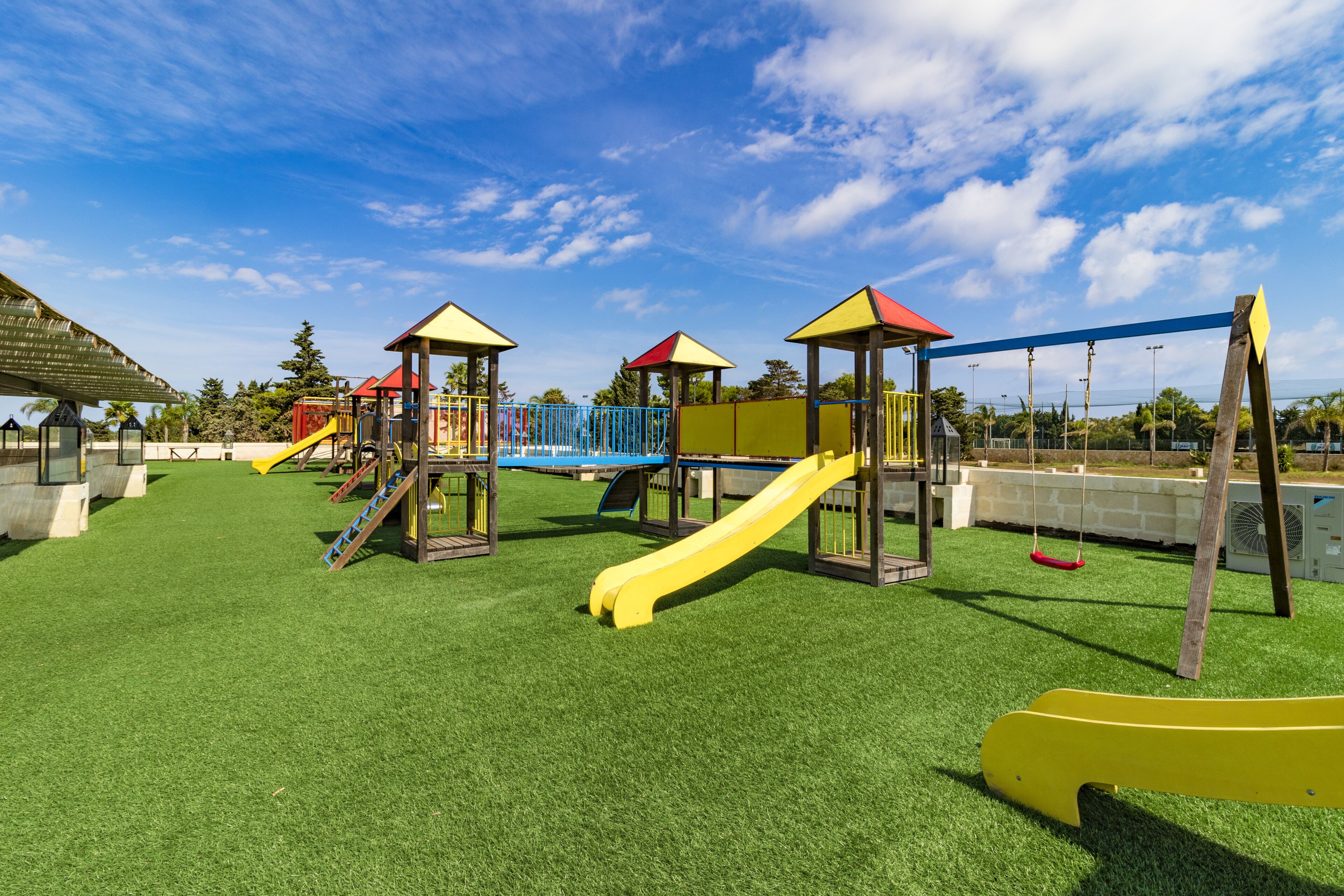 Ein Spielplatz mit mehreren Rutschen und Klettergerüsten, umgeben von einem Zaun und Rasen aus Kunststoff.