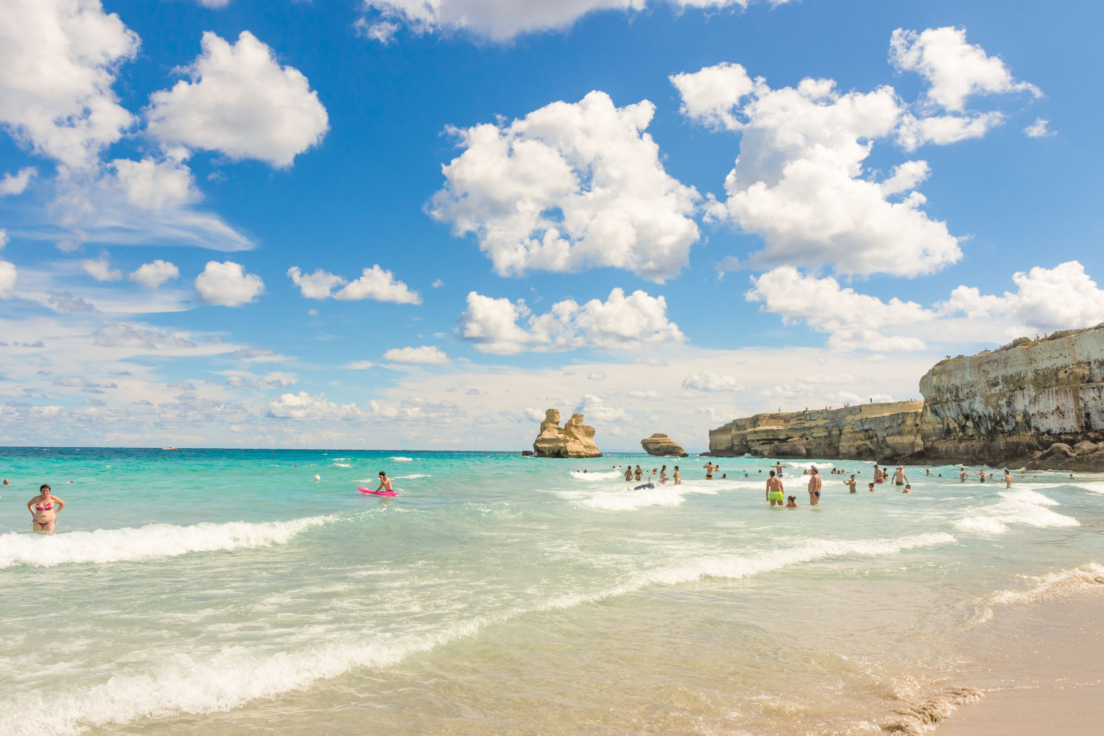 Una playa con aguas turquesas cristalinas, formaciones rocosas y gente nadando y relajándose.