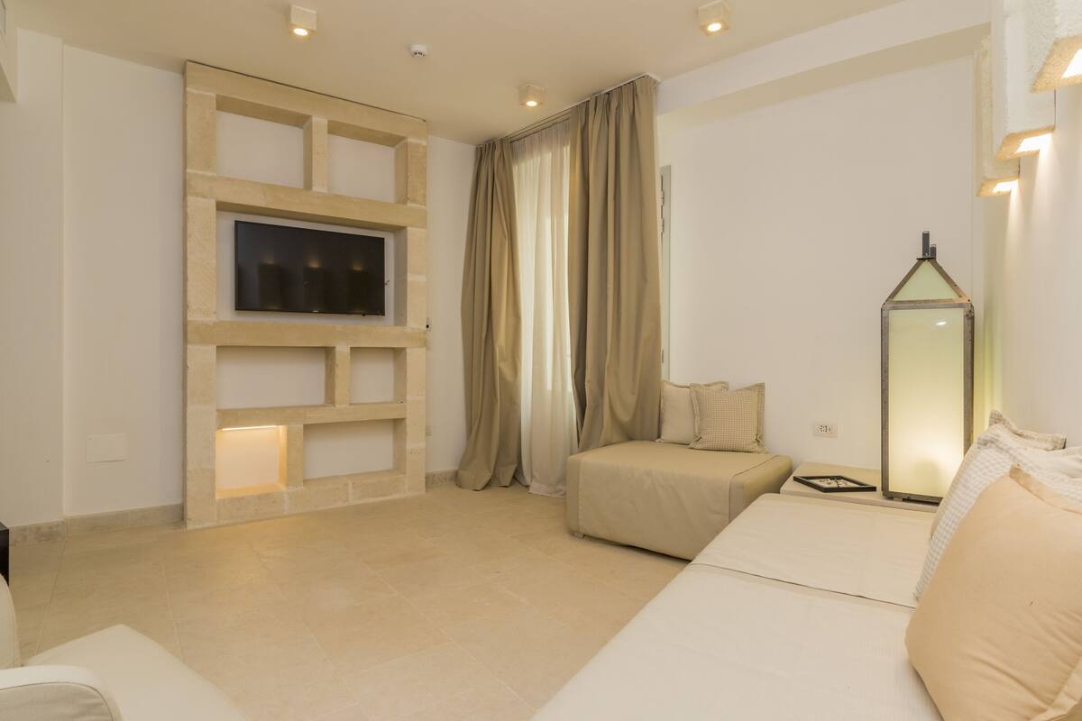Une chambre d’hôtel moderne dotée d’un téléviseur à écran plat fixé au mur, d’un lit avec une literie de couleur beige, d’un petit coin salon et d’une lampe de sol décorative.