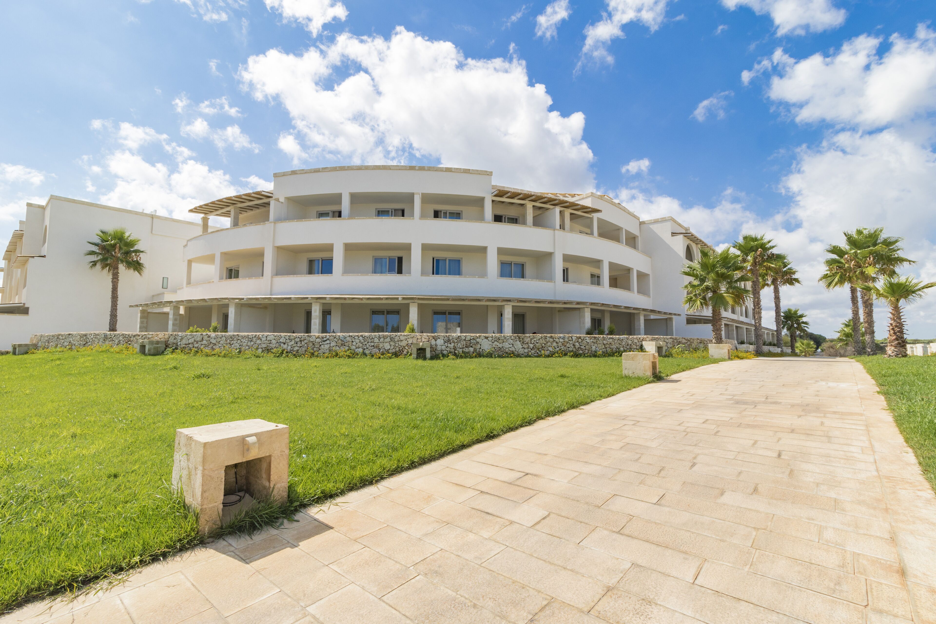 A modern multi-story building with a curved facade, a paved walkway, and palm trees.