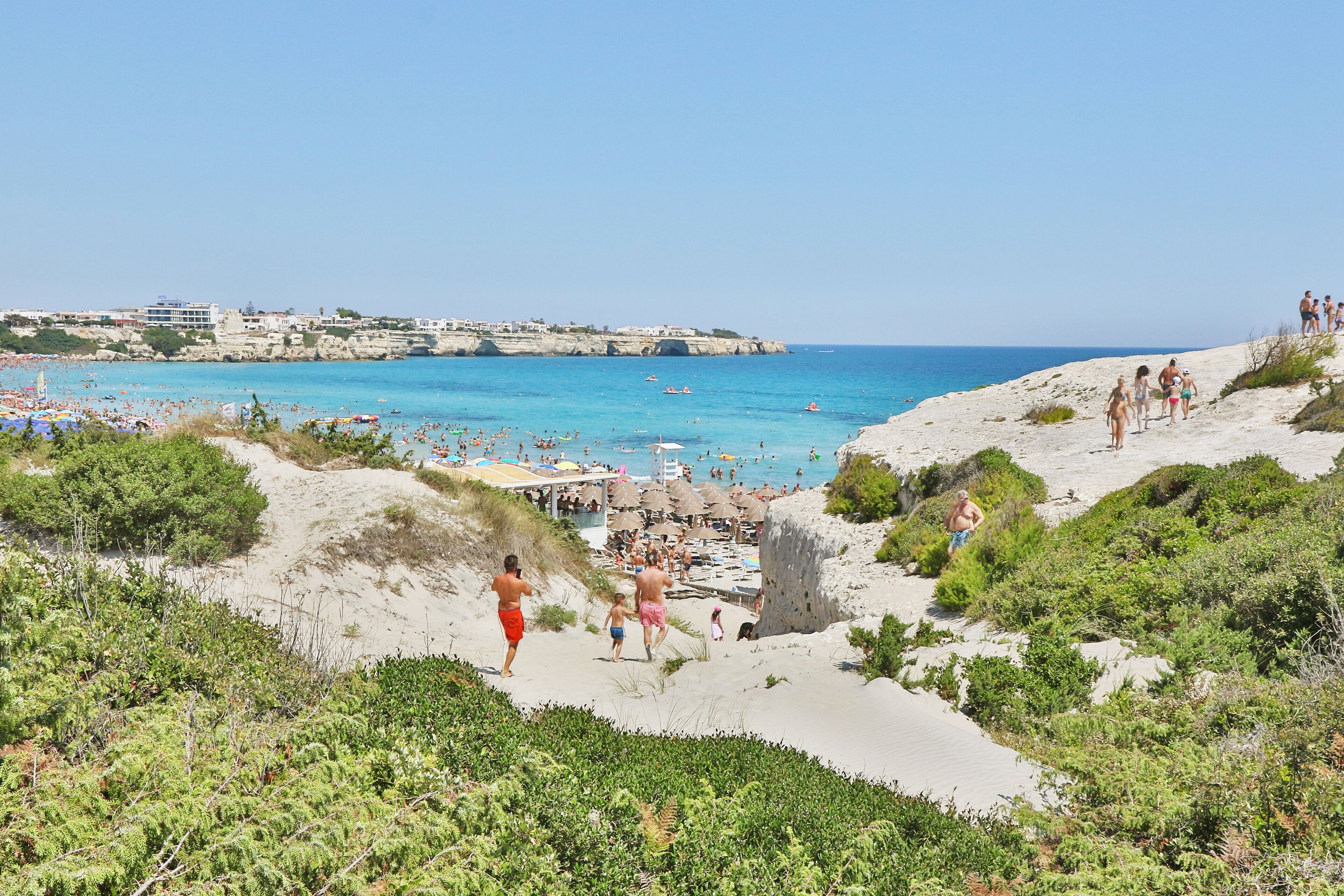 Une plage où l’on voit des gens se promener, un sentier de sable et, au loin, une vue sur la mer et les bâtiments.