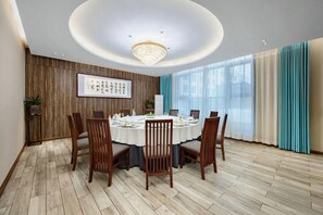 A dining room with a round table set for six, wooden chairs, a chandelier, a framed artwork on the wall, and large windows with curtains. - Botai Hotel (Yongqing)