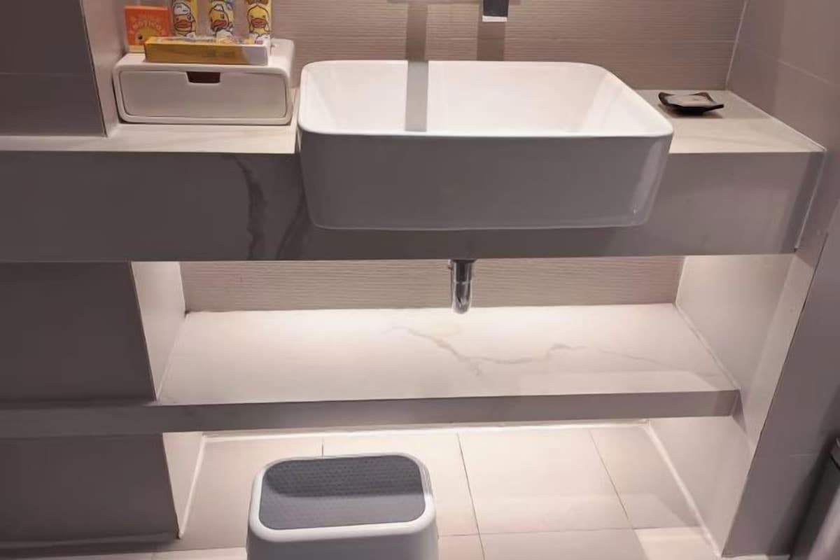 A modern bathroom with a rectangular sink, a small stool, and a shelf with toiletries.