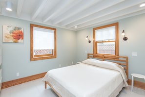 A bedroom with a bed, two windows with blinds, a wall-mounted lamp, and a small round table.