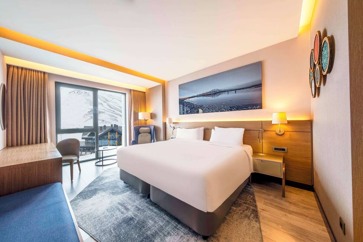Habitació d'hotel moderna amb un llit gran, zona de seients amb un sofà i una butaca, escriptori i una finestra gran amb vista a la ciutat.