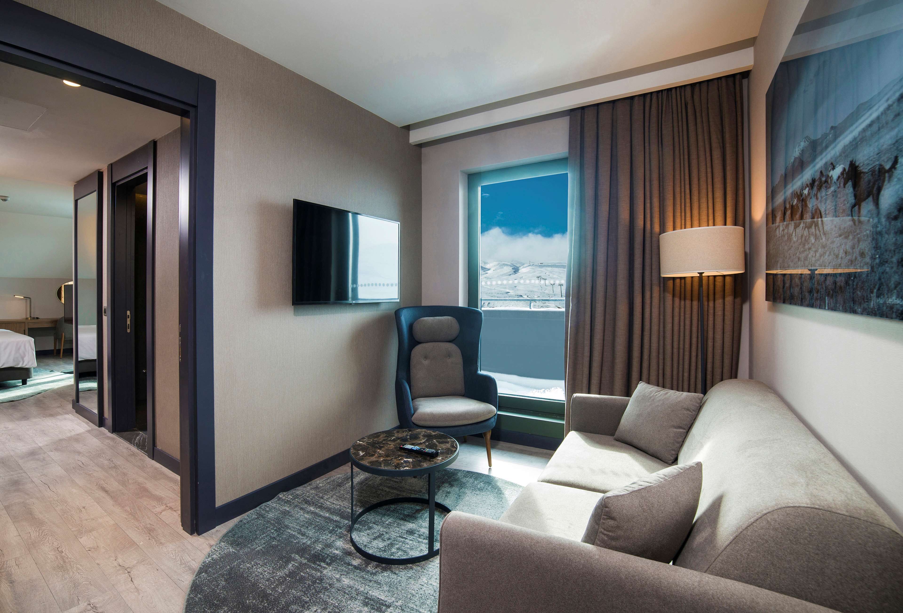 A hotel room with a sofa, a chair, a TV, a window with curtains, and a painting on the wall.