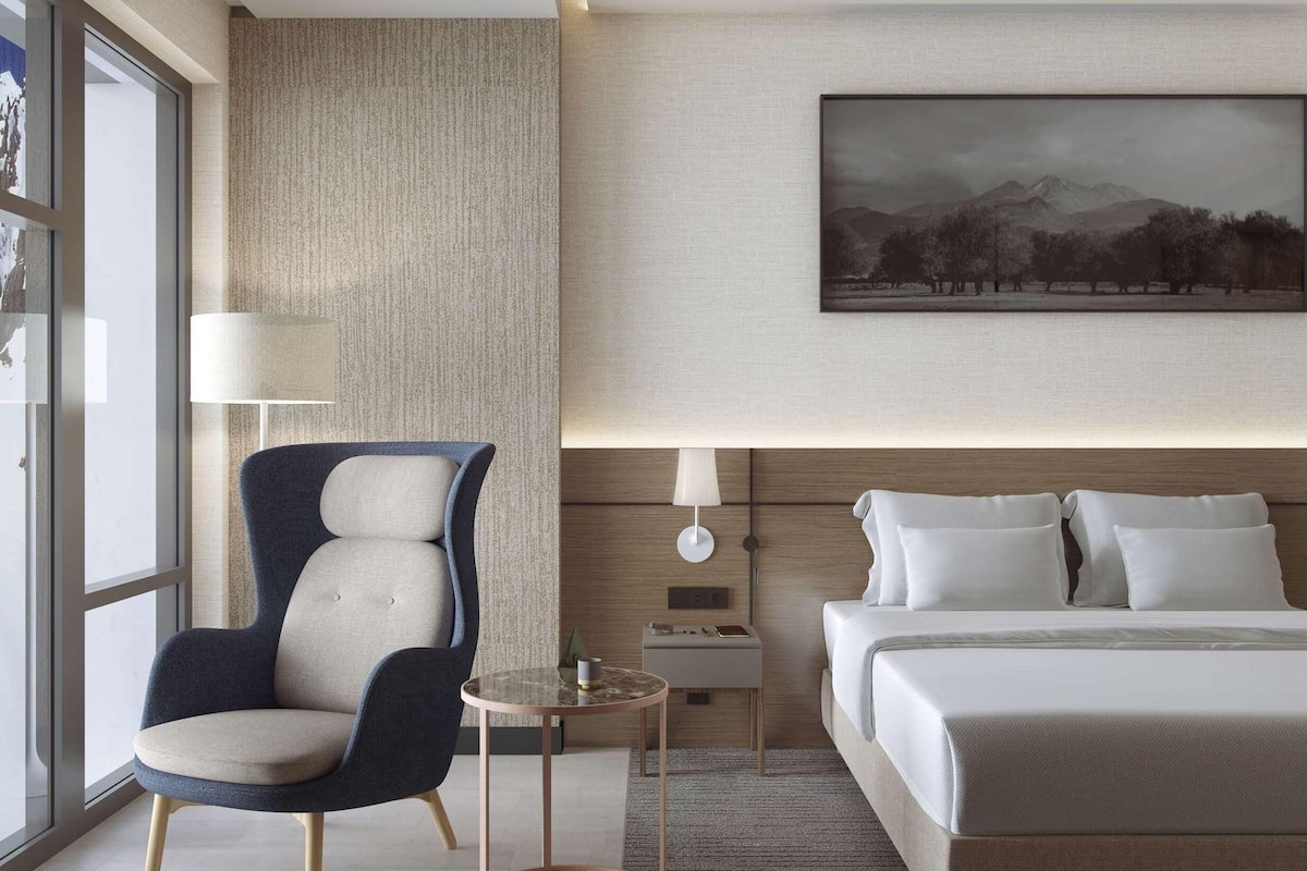Una habitació d'hotel moderna amb un llit gran, una zona d'estar amb una cadira i taula, i un finestral ample.