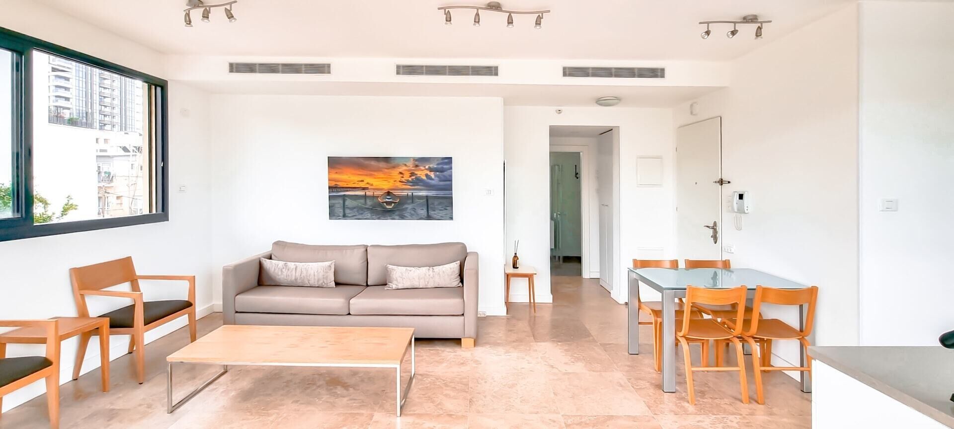 A modern living room with a beige sofa, wooden chairs, a glass dining table, and a painting of a sunset over water.