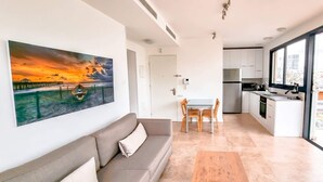 A modern apartment with a living area featuring a sofa and a painting, a dining area with a table and chairs, a kitchen with stainless steel appliances, and a large window offering a city view. - Apt Ophrys 1BR Tel Aviv Kalisher St TL66 (Tel Aviv)