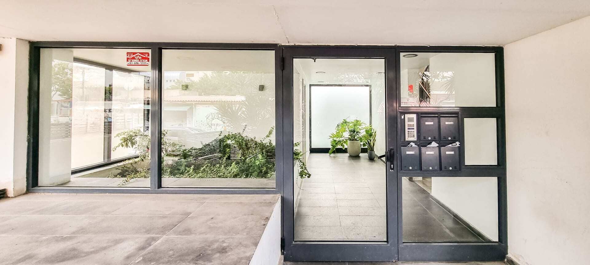 Entrada de un hotel con puertas de vidrio, un mostrador de recepción y una pequeña zona con plantas.