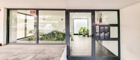 Entrada de un hotel con puertas de vidrio, un mostrador de recepción y una pequeña zona con plantas.