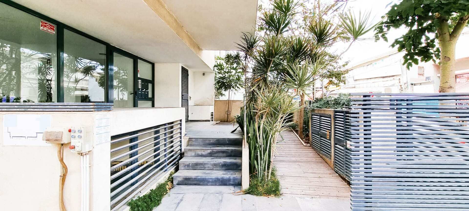 A narrow outdoor corridor with steps leading to a building entrance, flanked by metal fences and greenery.