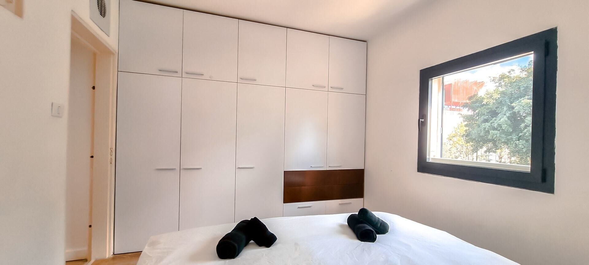 Une chambre à coucher comprenant un lit, une armoire et une fenêtre donnant sur des arbres et des immeubles.