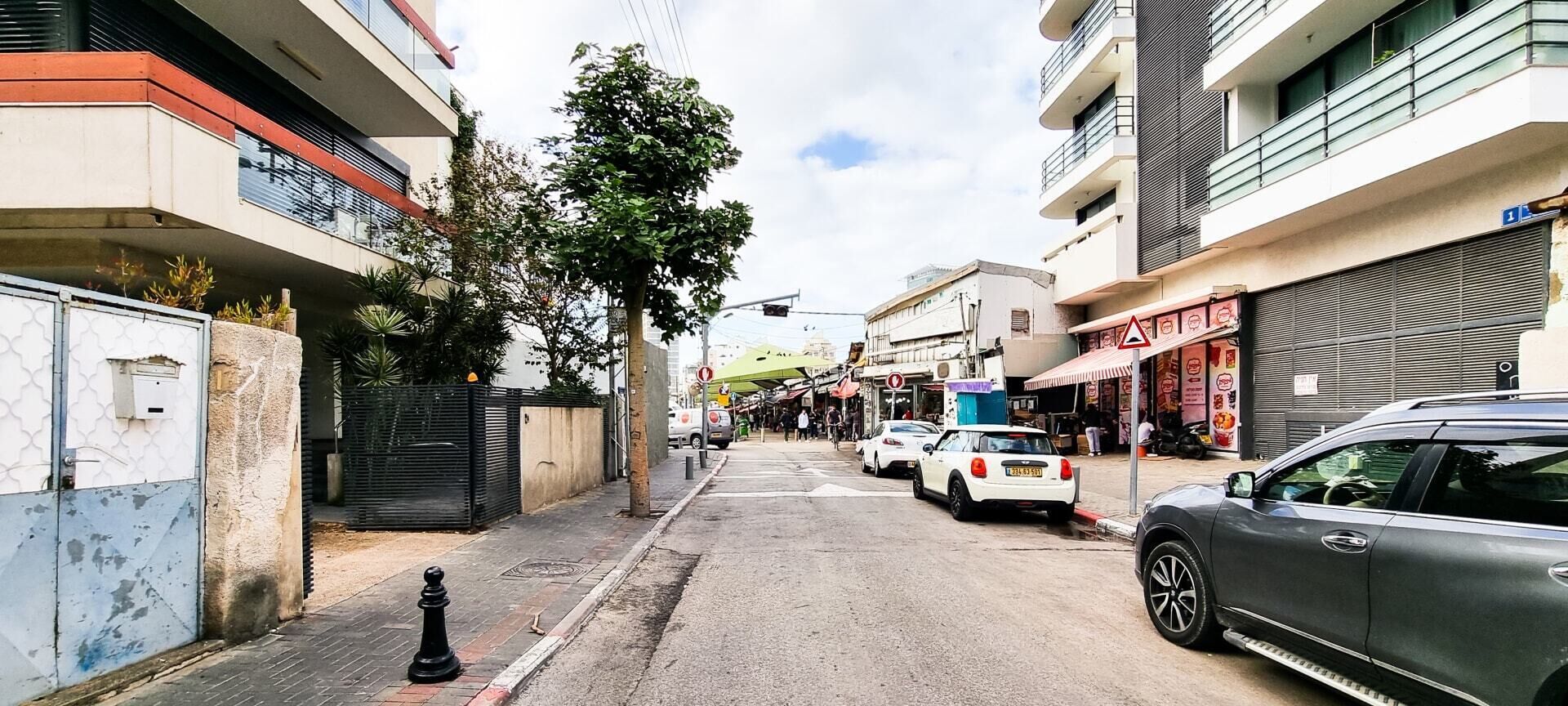Una stretta via cittadina fiancheggiata da edifici a più piani, auto parcheggiate e alcuni passanti.