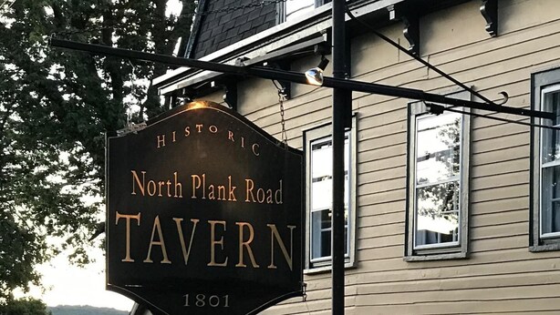 Uma taverna histórica com uma placa que diz "North Plank Road Tavern 1801" e uma bandeira americana.