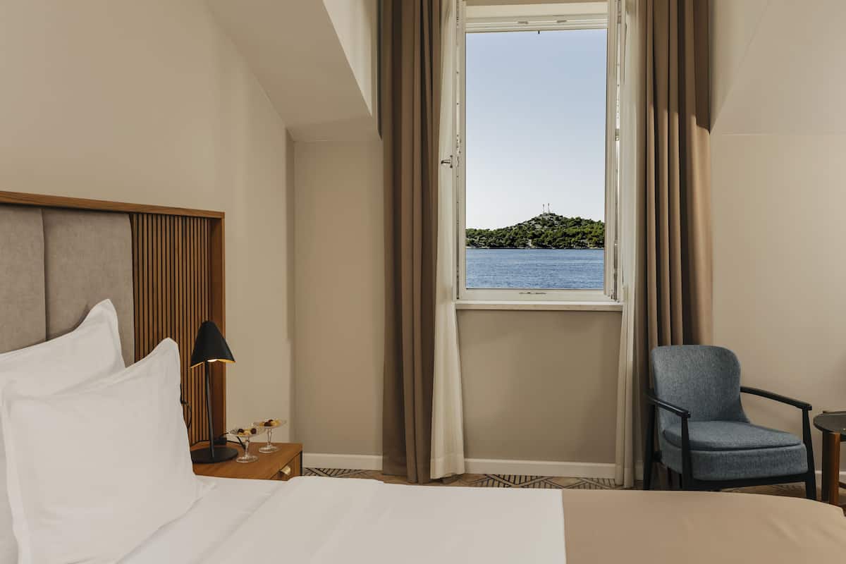 Une chambre d’hôtel comprenant un lit, une table de chevet, une lampe, une chaise et une fenêtre donnant sur un plan d’eau et une colline.
