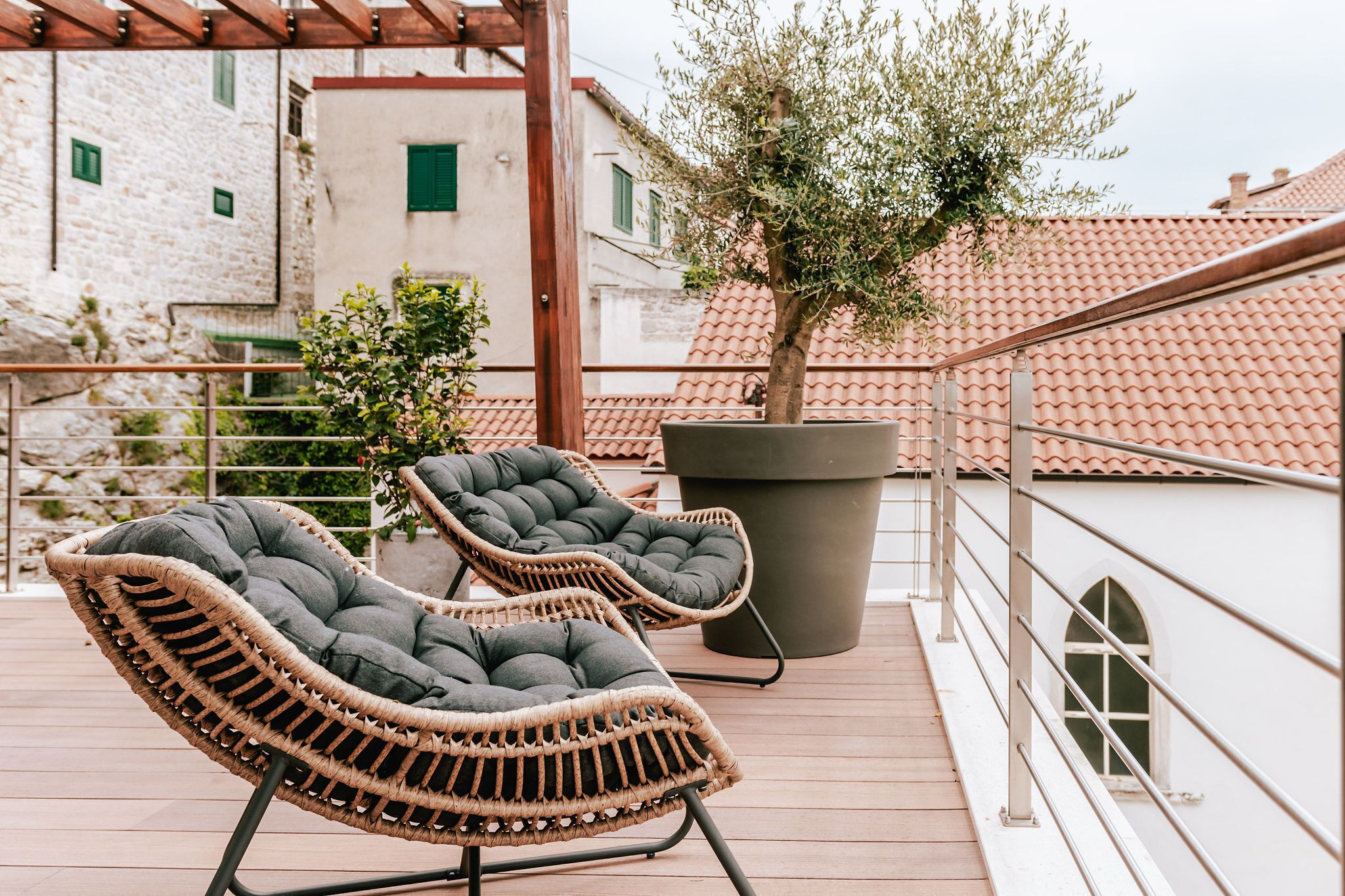 Dos sillones de mimbre con cojines en una terraza al aire libre con un olivo en maceta y vistas a edificios con tejados de tejas rojas.