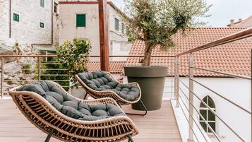 Dos sillones de mimbre con cojines en una terraza al aire libre con un olivo en maceta y vistas a edificios con tejados de tejas rojas.