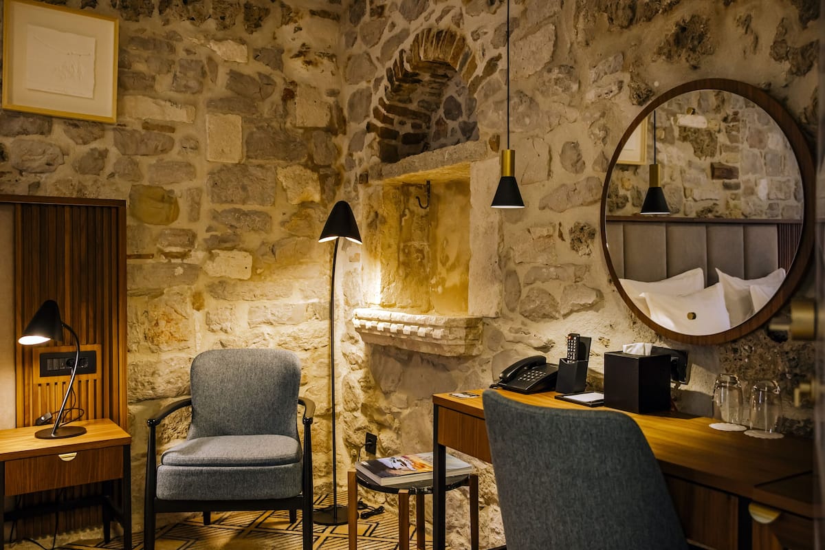 Une pièce avec un mur en pierre, un bureau en bois, deux chaises, une lampe sur pied, un téléphone et un miroir.