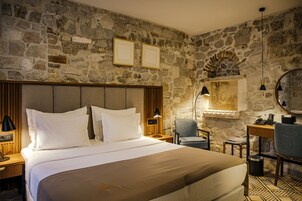 A hotel room with a stone wall, a bed with white bedding, a wooden headboard, a desk with a chair, and a small table with a lamp.