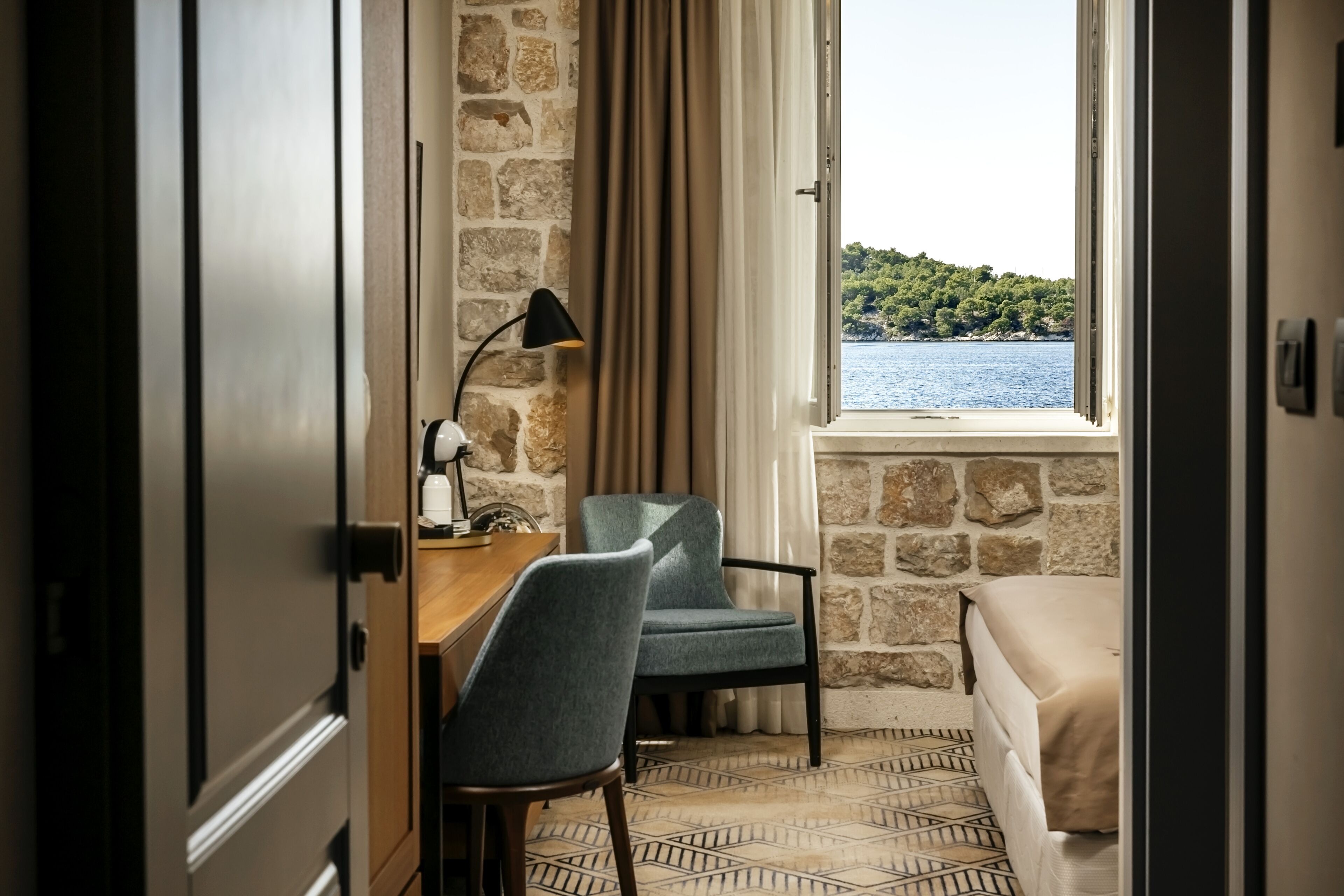 A room with a stone wall, a wooden desk with a lamp, a blue chair, a bed, and a view of a lake.