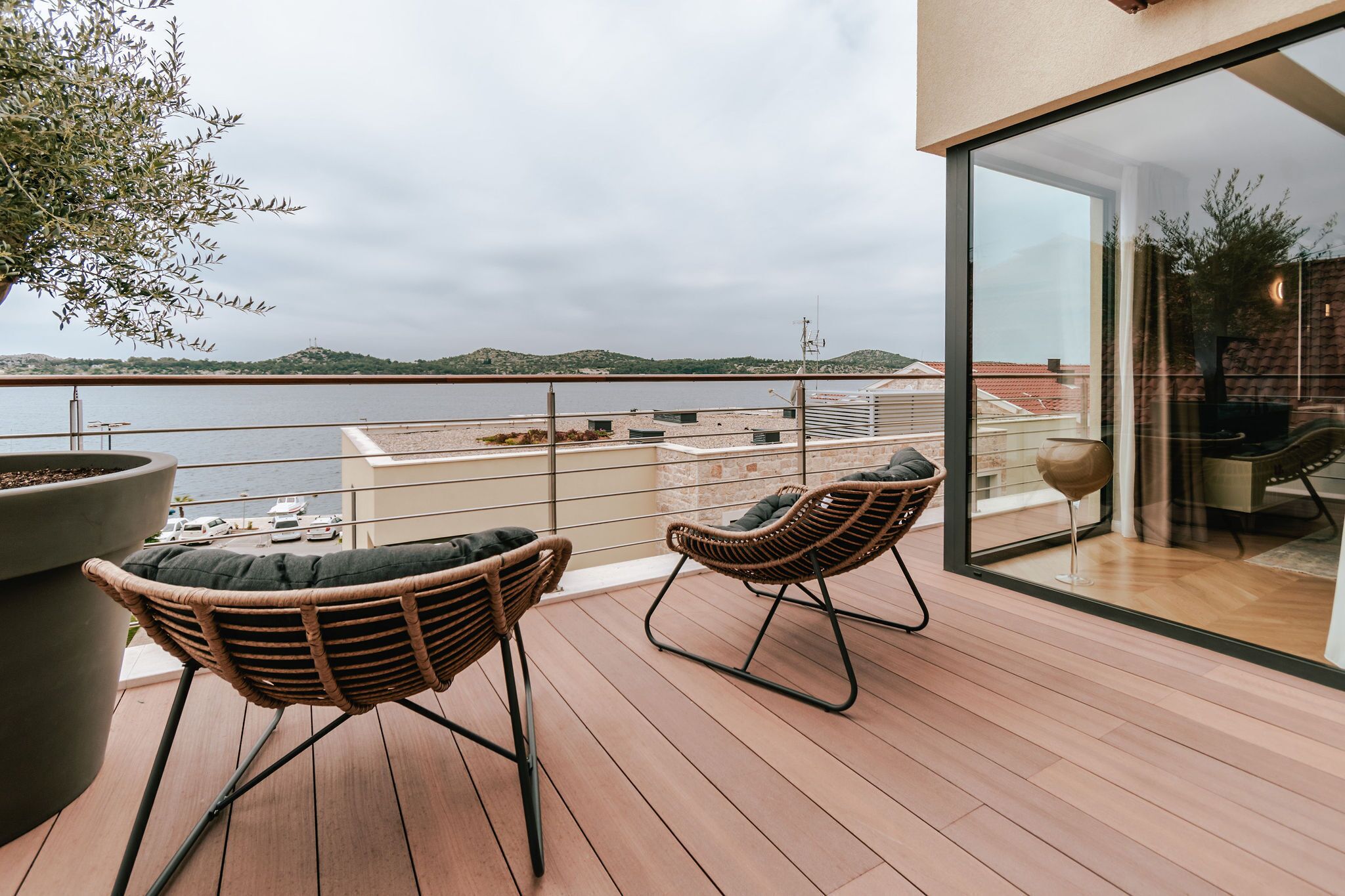 A balcony with wicker chairs, a large potted plant, and a view of the sea and distant hills.