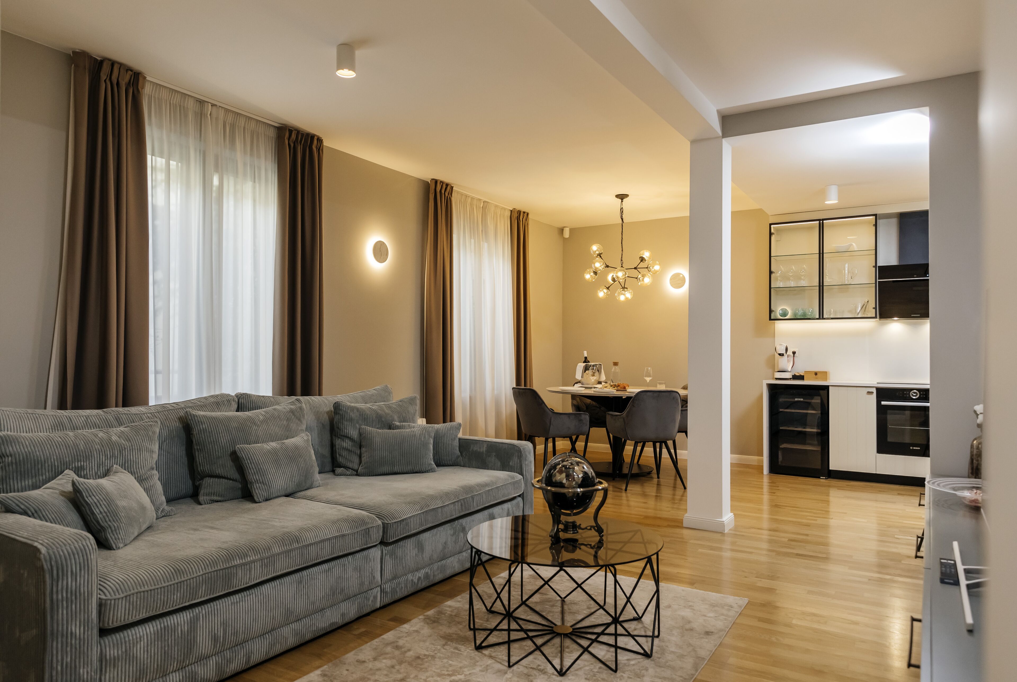 Una sala de estar moderna con un sofá gris, una mesa de comedor con sillas, una zona de cocina con armarios y electrodomésticos, y una araña.
