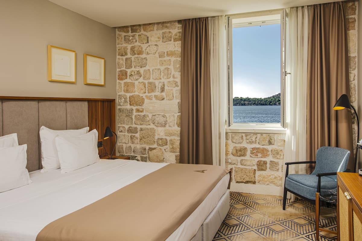 Une chambre d’hôtel avec un lit, une chaise, un bureau, une lampe, une fenêtre donnant sur la mer et des murs en pierre.