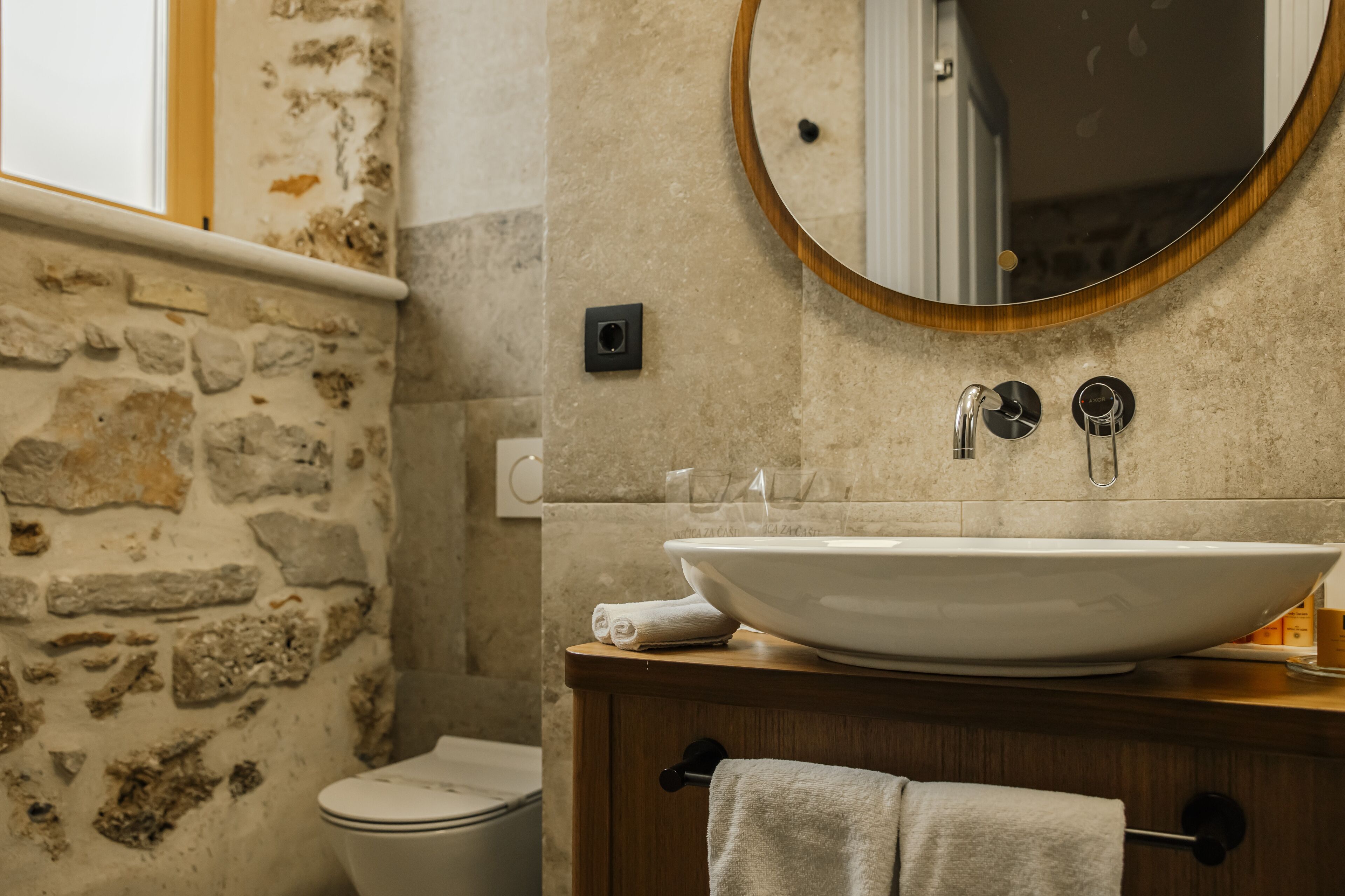 A bathroom with a stone wall, a wooden vanity, a white sink, a mirror, and a toilet.