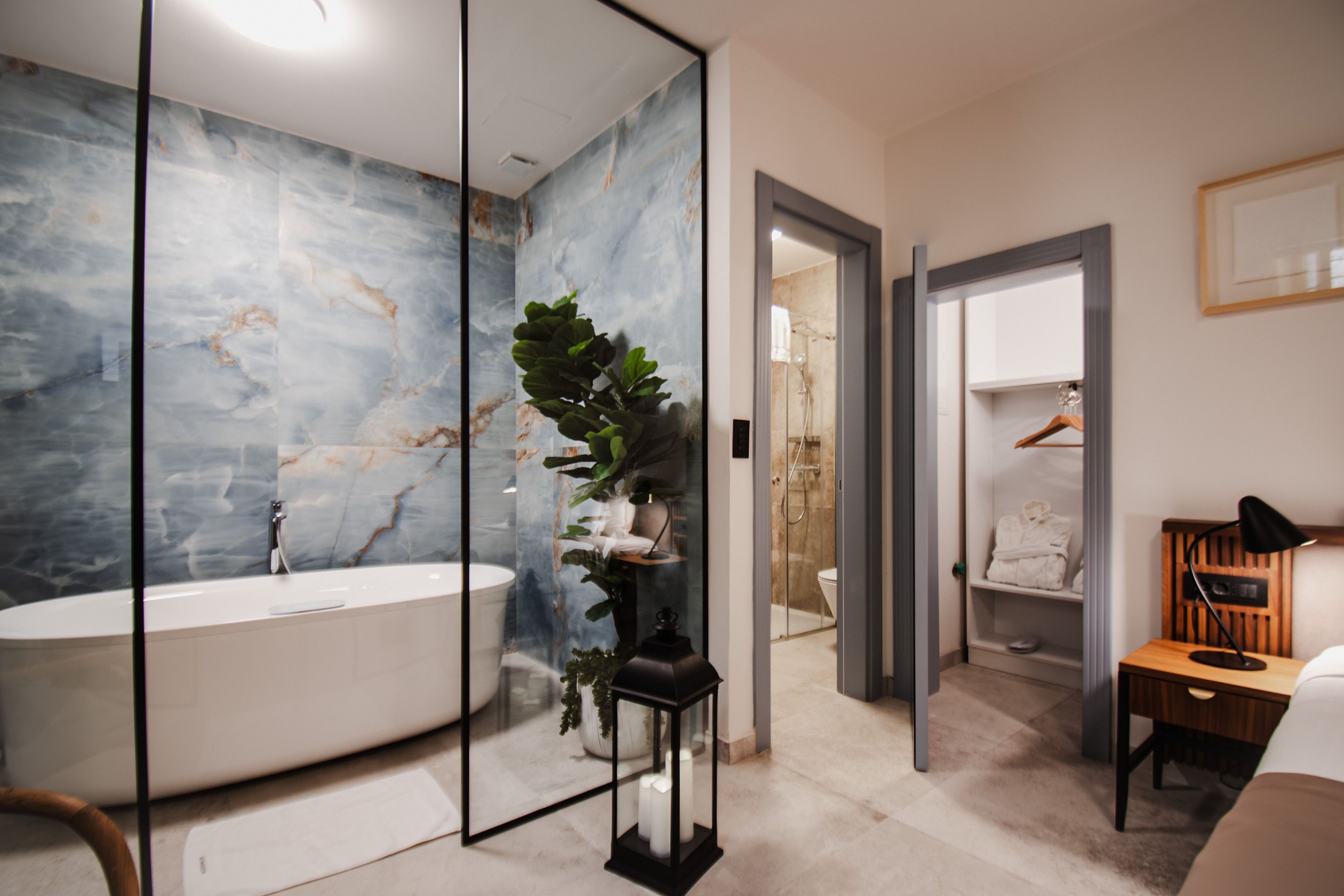 Un baño moderno con pared de mármol, una bañera independiente, una planta en maceta y una mesita de noche de madera con una lámpara.