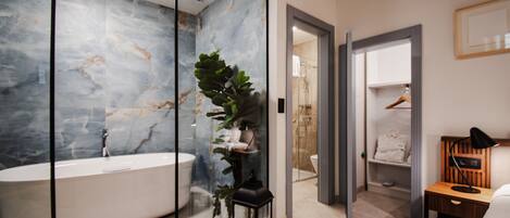 A modern bathroom with a marble wall, a freestanding bathtub, a potted plant, and a wooden bedside table with a lamp.